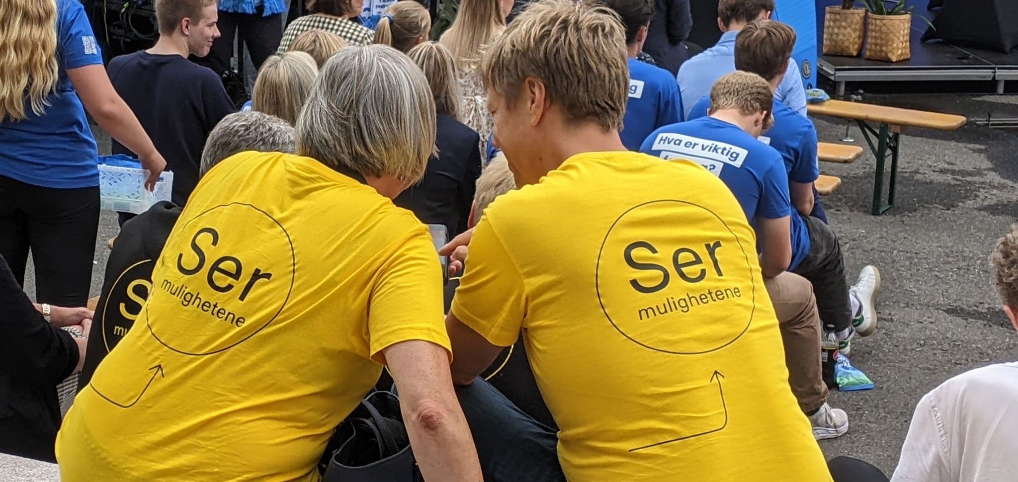To personer med gule t-skjorter med ryggen mot kamera. Tekst: Ser mulighetene.